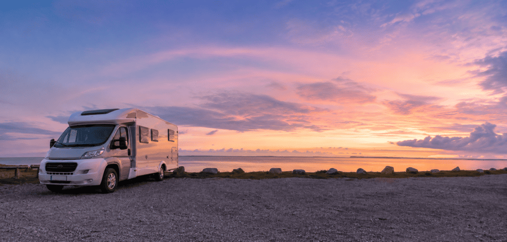 White motorhome parked on a gravel coastal overlook beside calm water under a vivid pink, orange, and purple sunset sky.